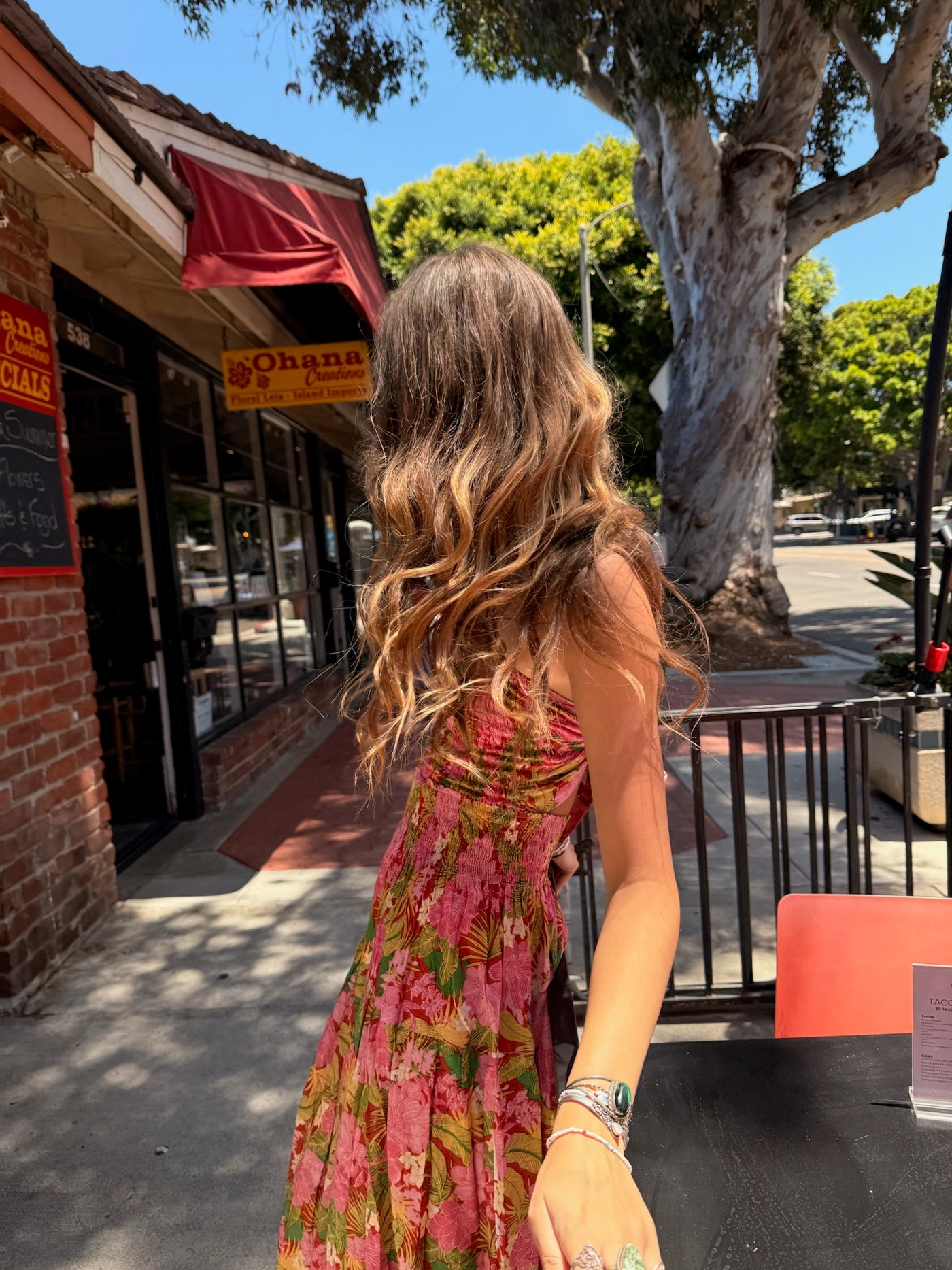 Woman in a pink dress standing on a sidewalk with a tree and storefronts in the background