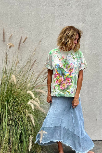 Woman wearing a floral t-shirt and denim skirt standing in front of a textured wall with tall grasses.