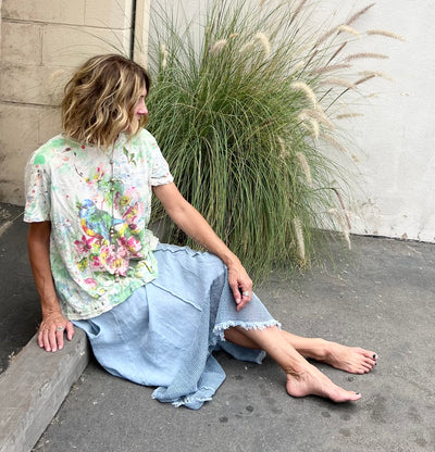 Woman sitting on a ledge wearing a floral shirt and blue skirt with tall grasses in the background.